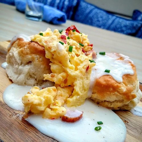 A fluffy biscuit topped with scrambled eggs, bacon, and creamy sauce, served on a wooden board.
