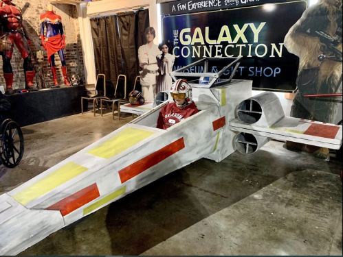 A child sits in a large, painted X-wing starfighter model at a themed shop, surrounded by various character displays.