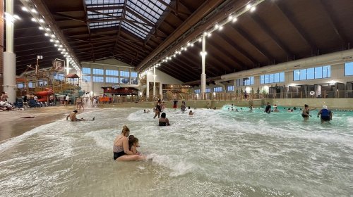 Indoor water park scene with people enjoying the wave pool and water slides under a high, wooden ceiling.