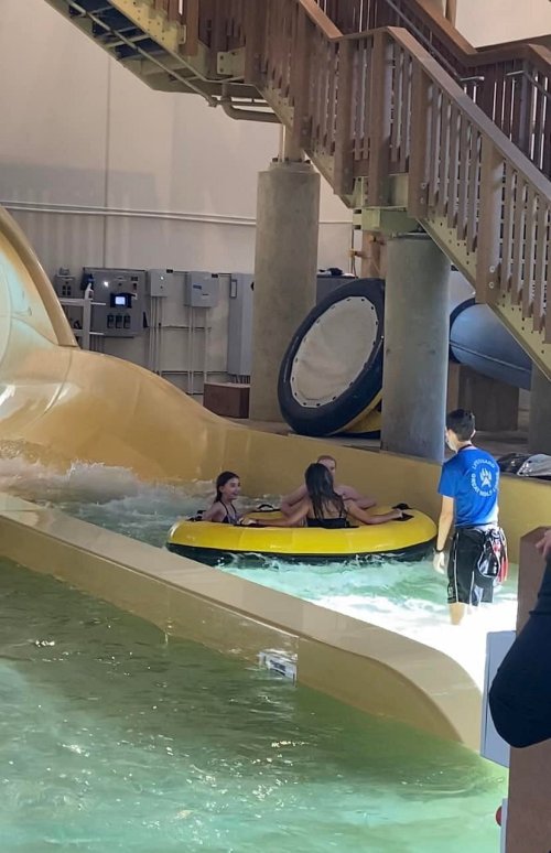 Two children on a yellow inflatable raft slide down a water slide, while a lifeguard watches nearby.