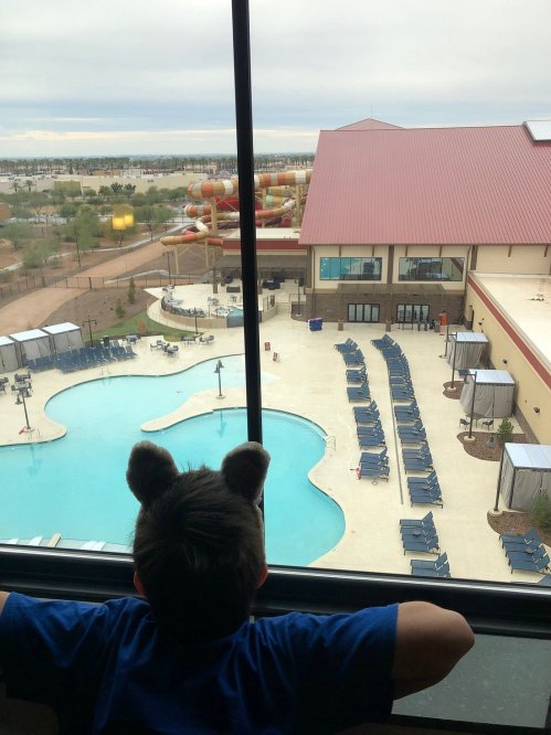 A child with bear ears looks out a hotel window at a pool and water slide area on a cloudy day.