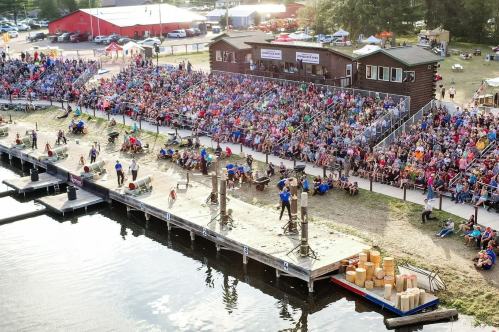 A large crowd gathers by a waterfront for an outdoor event, with performers on a stage and wooden structures nearby.