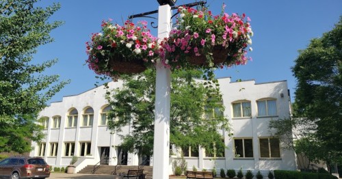 A white building with large windows, surrounded by green trees and colorful flower baskets hanging from a post.