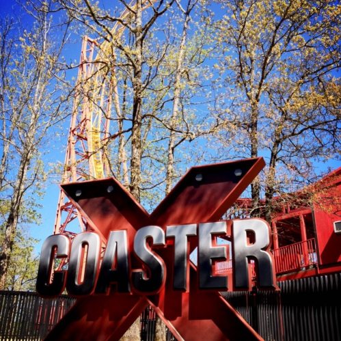 A large red "X" sign with the word "COASTER" in front of a tall amusement park ride and trees in the background.