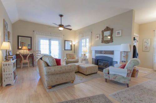 Cozy living room with a fireplace, two armchairs, an ottoman, and a dining area, featuring warm lighting and wooden floors.