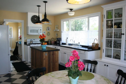Bright kitchen with black and white checkered floor, wooden island, and a table with flowers. Natural light from windows.