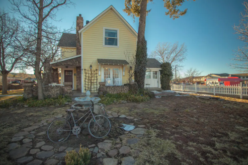 A charming yellow house with a stone path and a vintage bicycle in the foreground, surrounded by trees and a white fence.