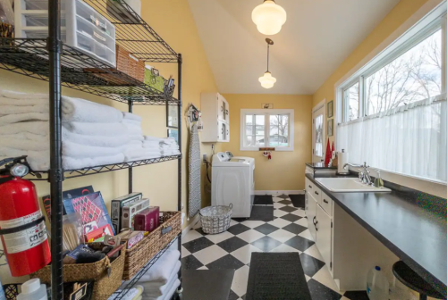 A bright laundry room with yellow walls, black and white checkered floor, shelves of towels, and a washing machine.