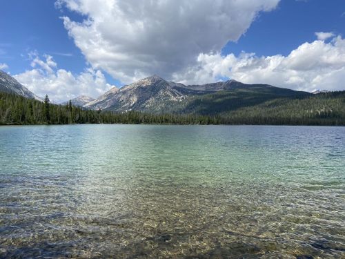 A serene lake surrounded by mountains under a partly cloudy sky, with clear water revealing pebbles beneath the surface.