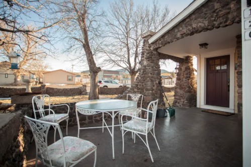 A stone porch with a round table and four white chairs, surrounded by trees and a view of nearby houses.
