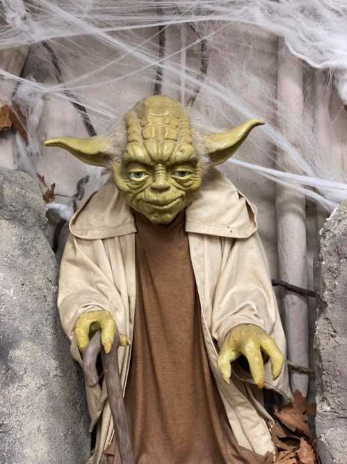 A lifelike Yoda figure with green skin and large ears, dressed in a brown robe, set against a spooky backdrop.