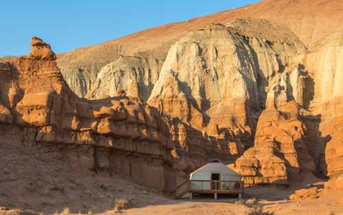 A yurt nestled among colorful rock formations in a desert landscape under a clear blue sky.