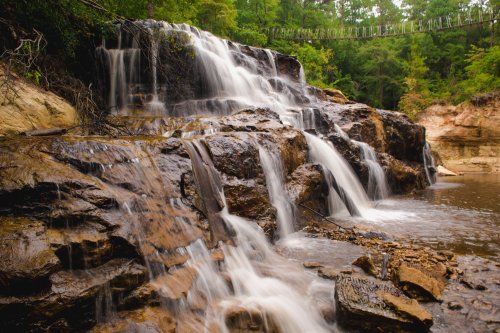 A serene waterfall cascades over rocky terrain, surrounded by lush green trees and a peaceful river below.