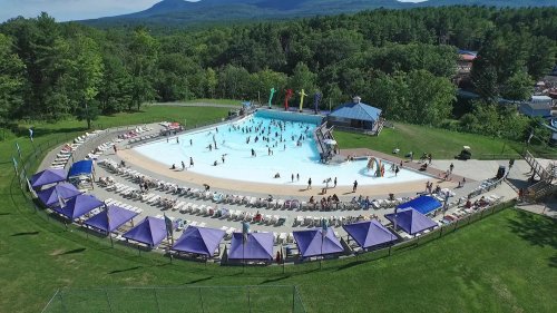 Aerial view of a large swimming pool surrounded by green trees, with people swimming and lounging on deck chairs.