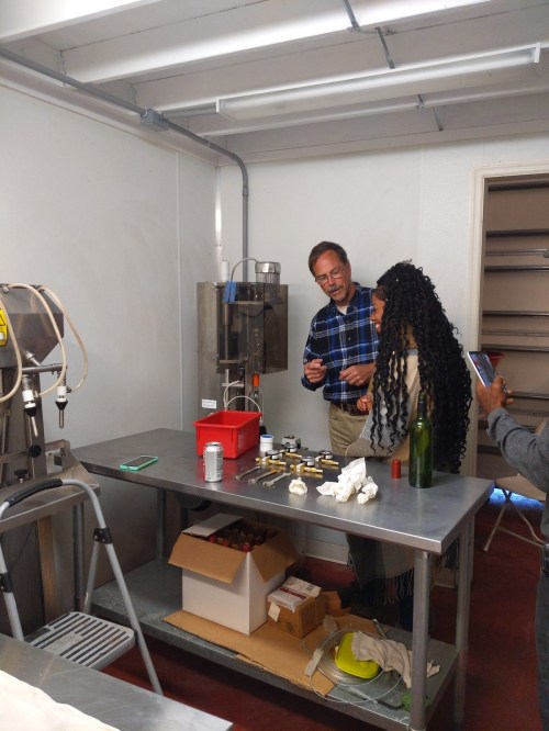 A man and a woman discuss equipment in a lab setting, with various tools and a bottle on a stainless steel table.