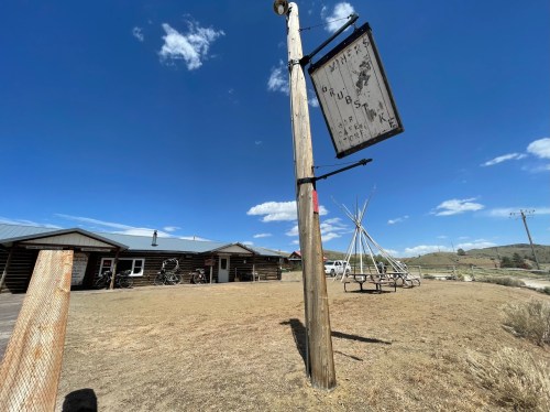 A rustic building with a wooden sign, surrounded by open land and a teepee, under a clear blue sky.