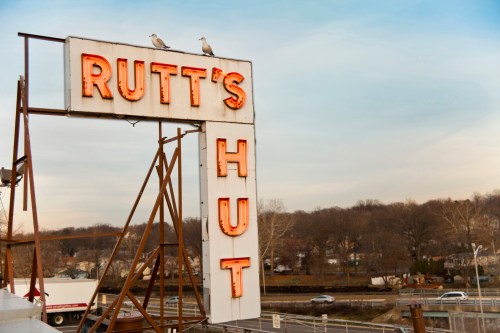 Neon sign for Rutt's Hut with two seagulls perched on top, set against a cloudy sky and distant trees.