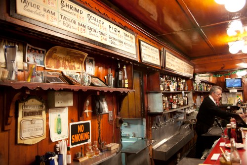 A vintage bar with wooden paneling, a menu board, and a bartender preparing drinks. "No Smoking" sign visible.