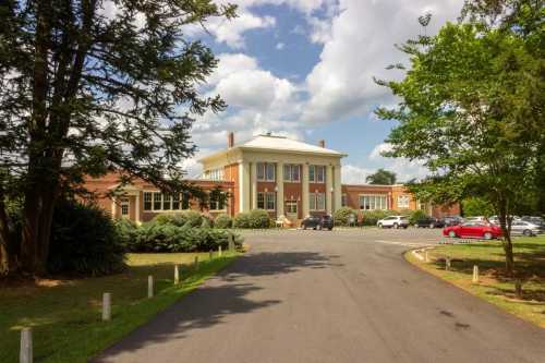 A large brick building with a white portico, surrounded by trees and a paved driveway, under a partly cloudy sky.