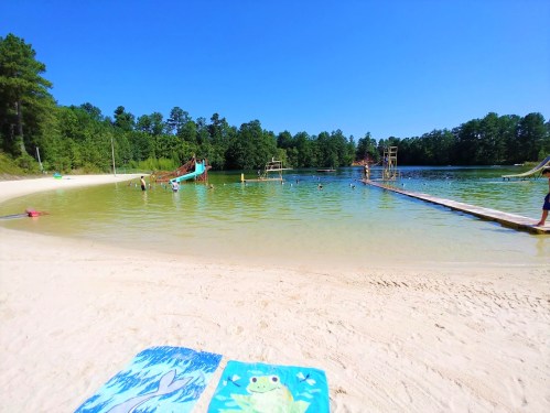 A sunny beach scene with a sandy shore, water slides, and people swimming in a clear lake surrounded by trees.