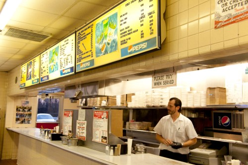 A diner interior with a menu board, a cashier in a white shirt, and food preparation area.