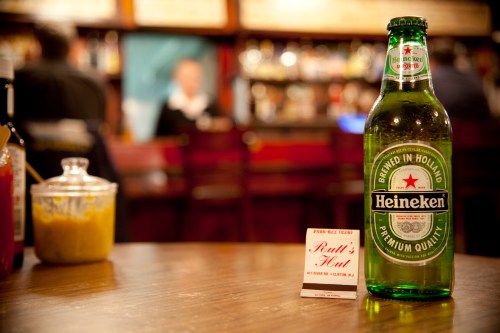 A bottle of Heineken beer on a wooden table with a business card for "Paul's Hut" in a bar setting.
