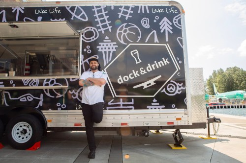 A chef stands beside a food truck with a "dock & drink" logo, featuring playful lake-themed graphics in the background.