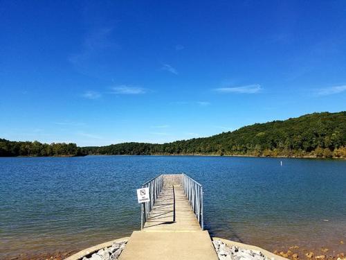 A serene lake view with a wooden dock extending into the water, surrounded by green hills under a clear blue sky.
