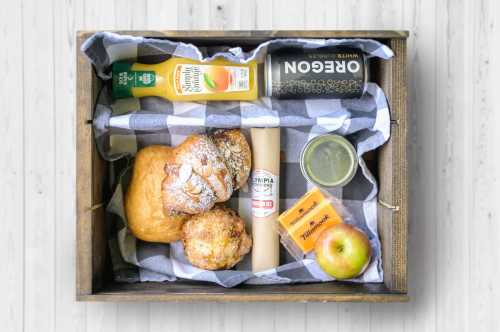 A wooden crate filled with breakfast items: pastries, juice, a can, an apple, and a roll, all on a checkered cloth.