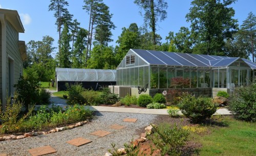 A sunny garden scene featuring greenhouses, a gravel path, and lush greenery surrounded by trees.