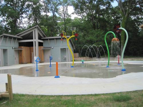 Colorful water spray features in a splash pad area, surrounded by trees and a building in the background.