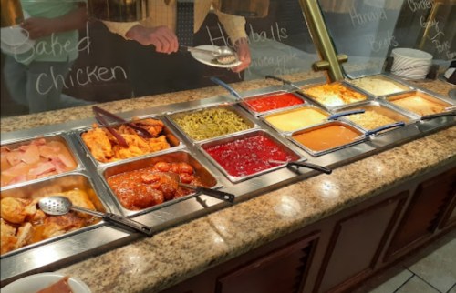 A buffet with various dishes including baked chicken, meatballs, and sides in metal trays on a granite countertop.