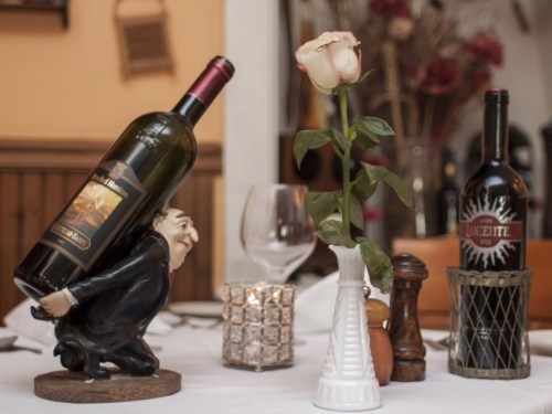 A whimsical wine holder with a bottle, a rose in a vase, and two wine bottles on a dining table setting.