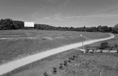 A wide, empty field with a blank billboard and a gravel path, surrounded by trees under a clear sky.