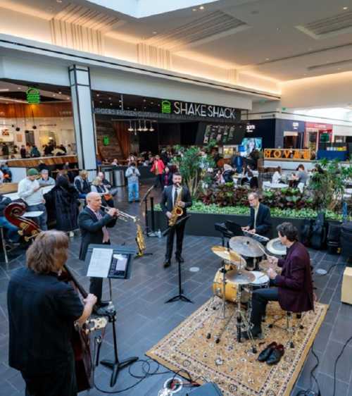 A live jazz band performs in a bustling mall, surrounded by greenery and shoppers enjoying the atmosphere.