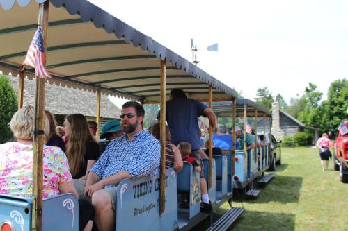 A group of people sitting on a train ride in a park, with flags and greenery in the background.