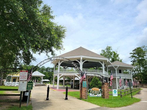 Entrance to a farmers market in a park, featuring a large gazebo and decorative signage, surrounded by greenery.