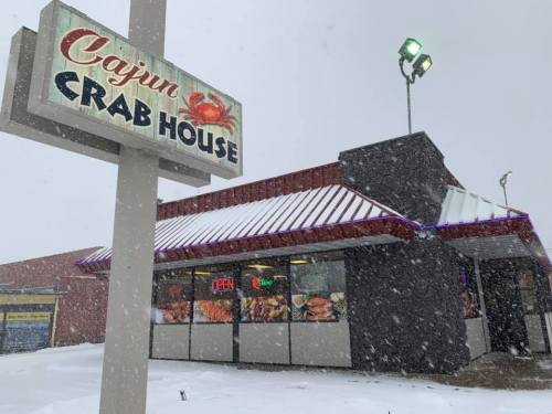 A snow-covered Cajun Crab House restaurant with a lit "OPEN" sign and a crab-themed sign outside.