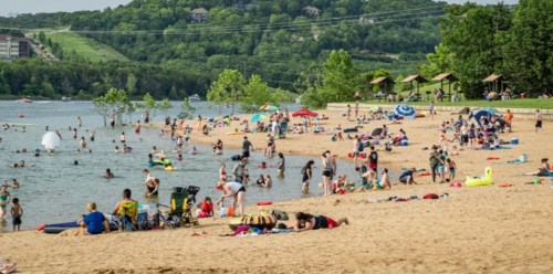 A busy beach scene with people swimming, sunbathing, and relaxing on the sand near a lake surrounded by greenery.