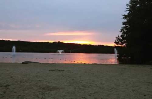 Sunset over a calm lake with fountains, surrounded by trees and sandy shore, under a cloudy sky.