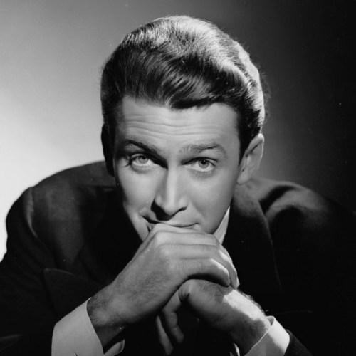 A black and white portrait of a man with slicked-back hair, smiling and resting his chin on his hands.