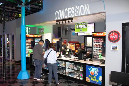A concession stand with various drinks and snacks, featuring customers placing orders at the counter.