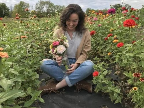 A person sits in a flower field, smiling and holding a bouquet of colorful flowers, surrounded by vibrant blooms.