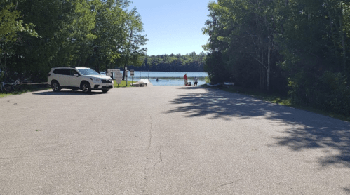 A serene lakeside view with a parking area, trees, and a person near the water on a sunny day.