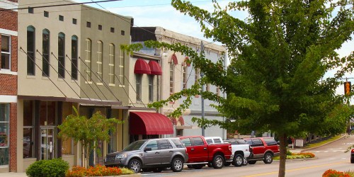 A street view of a small town with shops, parked trucks, and a tree lining the sidewalk on a sunny day.