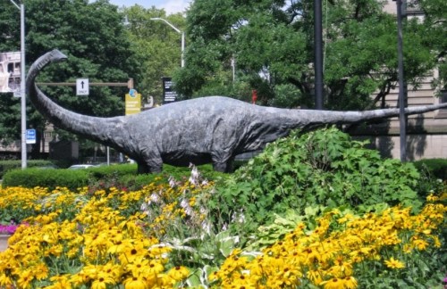 A large dinosaur statue surrounded by vibrant yellow flowers in a park setting.