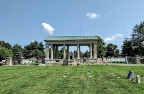 A stone mausoleum with a green roof surrounded by a grassy area and gravestones under a clear blue sky.