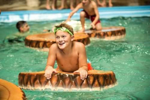 A smiling boy in swim trunks plays on a floating log in a pool, surrounded by other children enjoying the water.