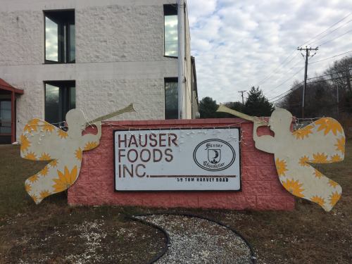 Sign for Hauser Foods Inc. featuring two decorative angels with trumpets and floral designs, set against a cloudy sky.