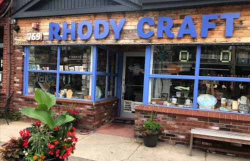 Exterior of Rhody Craft store with blue trim, wooden signage, and colorful flower planters in front.